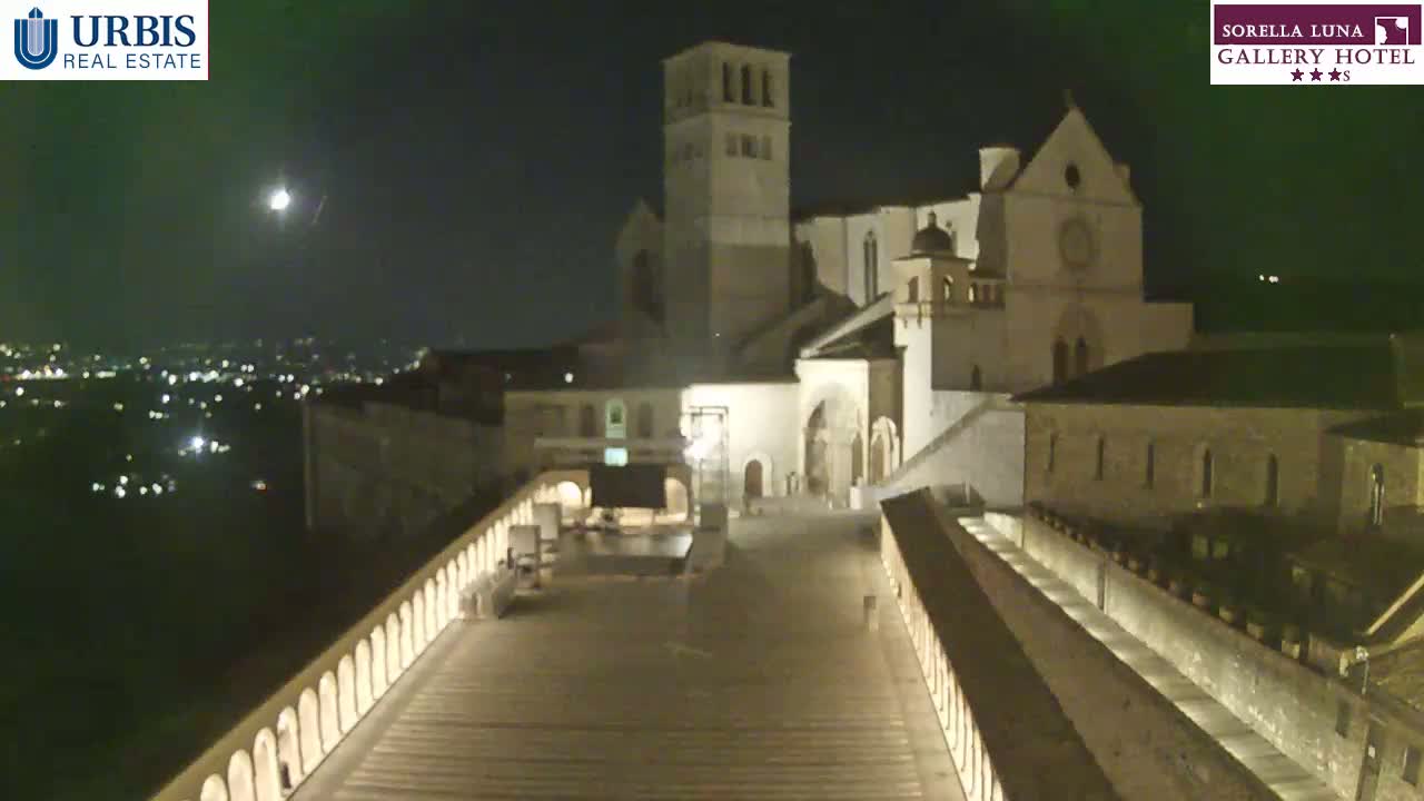 A nighttime view of a large stone building complex, possibly a church or monastery, illuminated under a clear, moonlit sky overlooking a city in the distance.