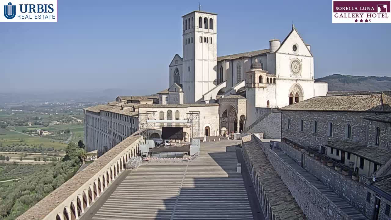 A large, light-colored basilica and adjoining buildings sit atop a hill on a clear, sunny day, overlooking a valley.