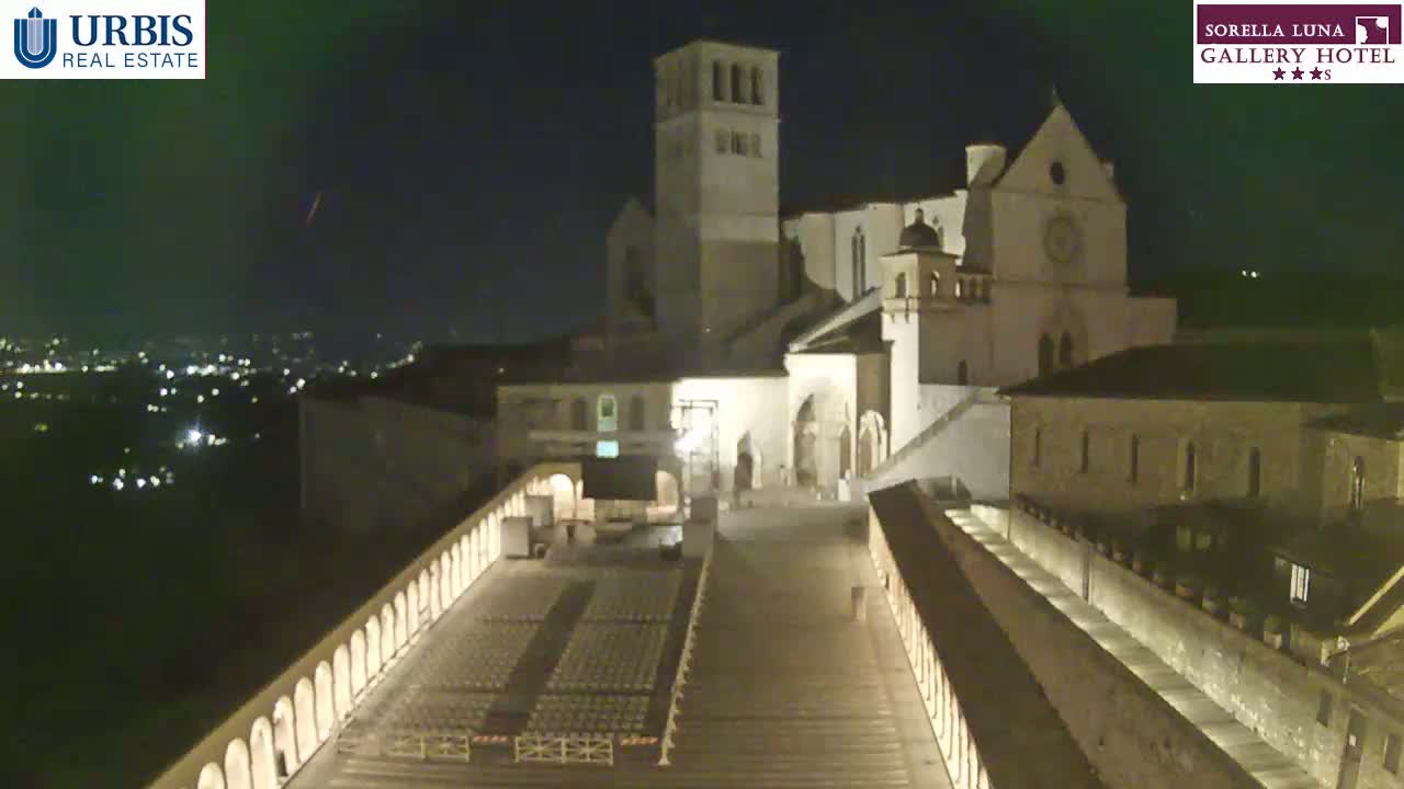 A large, illuminated stone building, possibly a church, sits on a hill at night, overlooking a city with many lights; a wide, long staircase leads down to a plaza below.