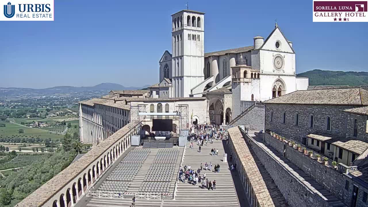 A large, light-colored basilica sits atop a hill overlooking a sprawling landscape under a clear, sunny sky; a large set of stairs leads down to a plaza below, where many people are gathered.