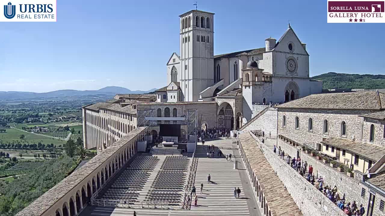 A large stone basilica and adjoining buildings are situated on a hill overlooking a valley on a sunny day, with a crowd of people gathered in the plaza below.