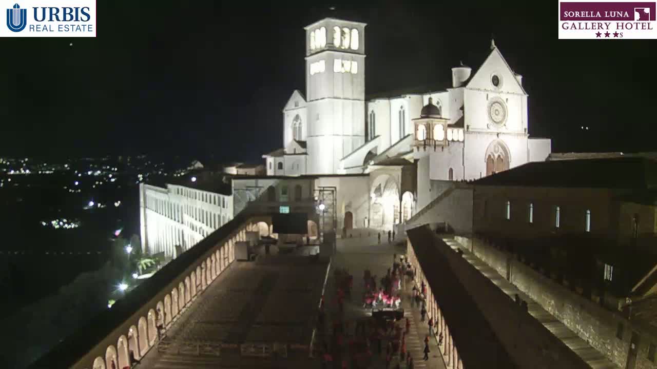 A large, illuminated basilica stands at night, overlooking a plaza with people gathered below, surrounded by low stone walls.