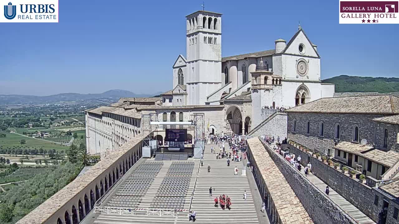 A large stone basilica and adjoining buildings are viewed from above on a sunny day, with a crowd of people gathered in the foreground on a large set of stairs leading to the church.