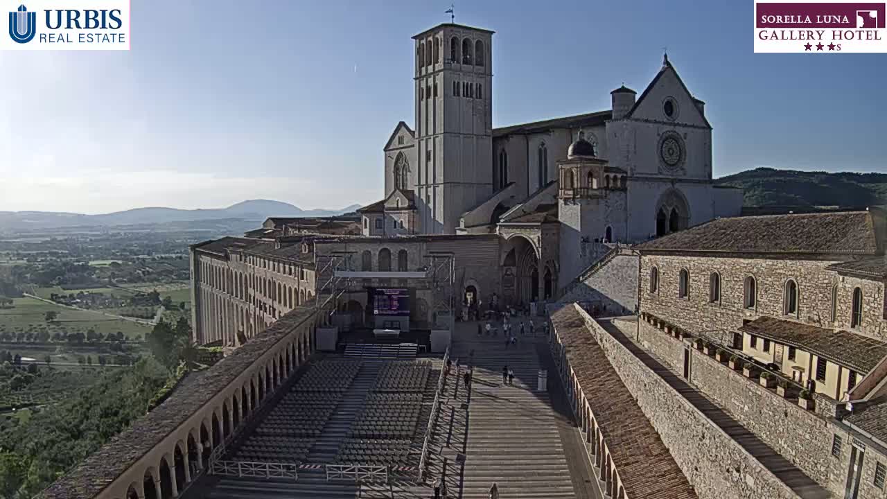 A large stone basilica and adjoining buildings overlook a wide plaza with empty seating, all under a clear, sunny sky.