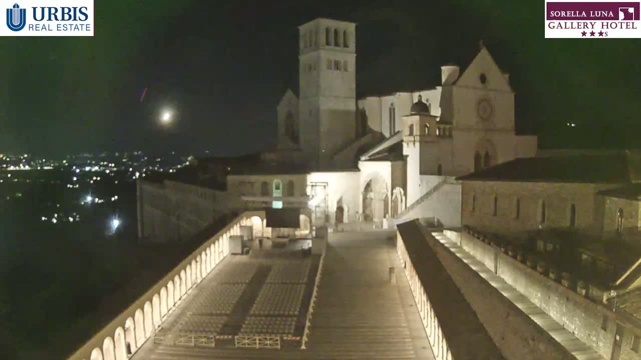 A large, illuminated basilica stands at night, overlooking a city with an empty plaza containing rows of chairs in the foreground under a partly cloudy sky.
