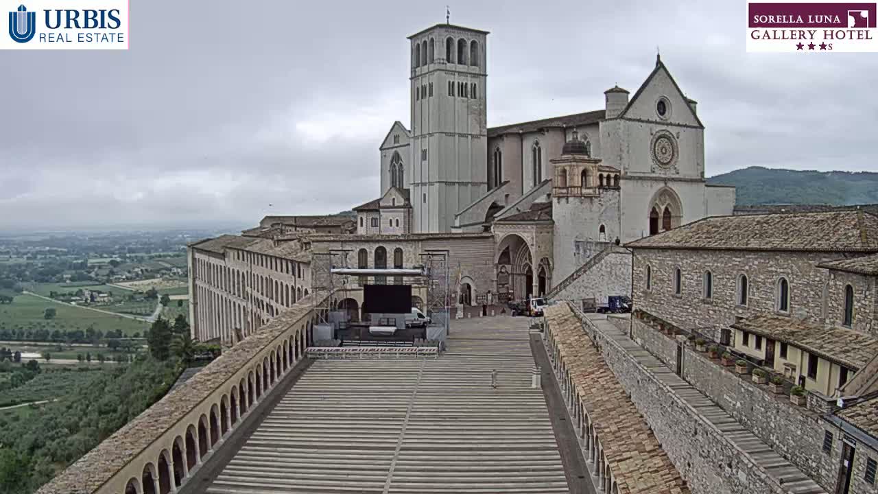 A large stone basilica and adjoining buildings are seen on a hilltop overlooking a verdant valley under an overcast sky.