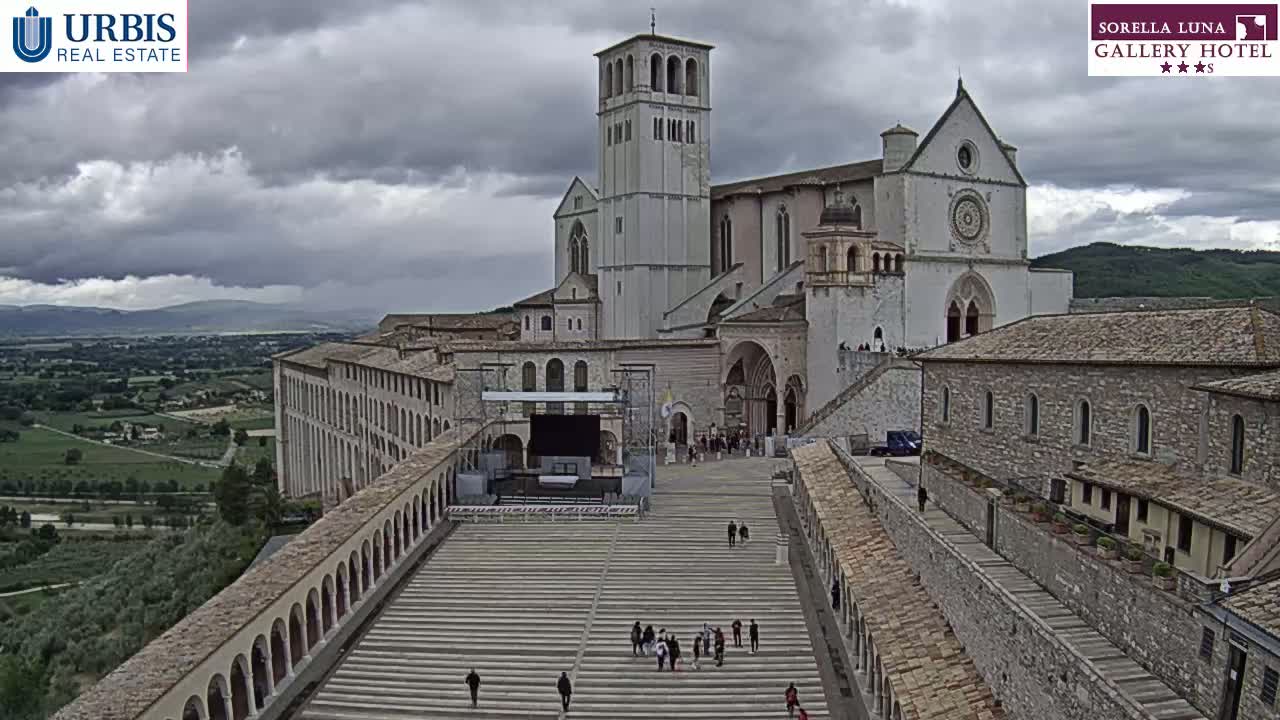 A large stone basilica and adjacent buildings are seen on an overcast day, with a wide staircase leading down to a plaza in the foreground, surrounded by low stone walls.