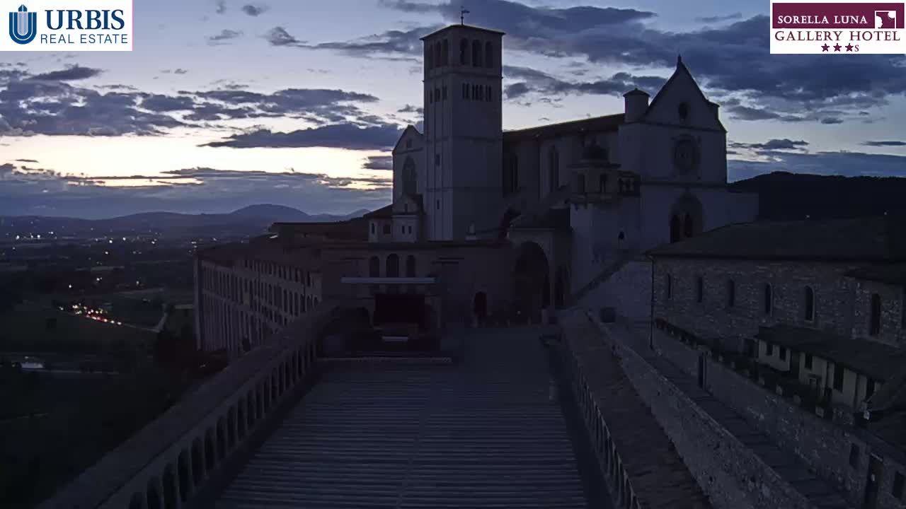 A large stone basilica sits on a hilltop at dusk, overlooking a valley filled with lights, under a cloudy sky.