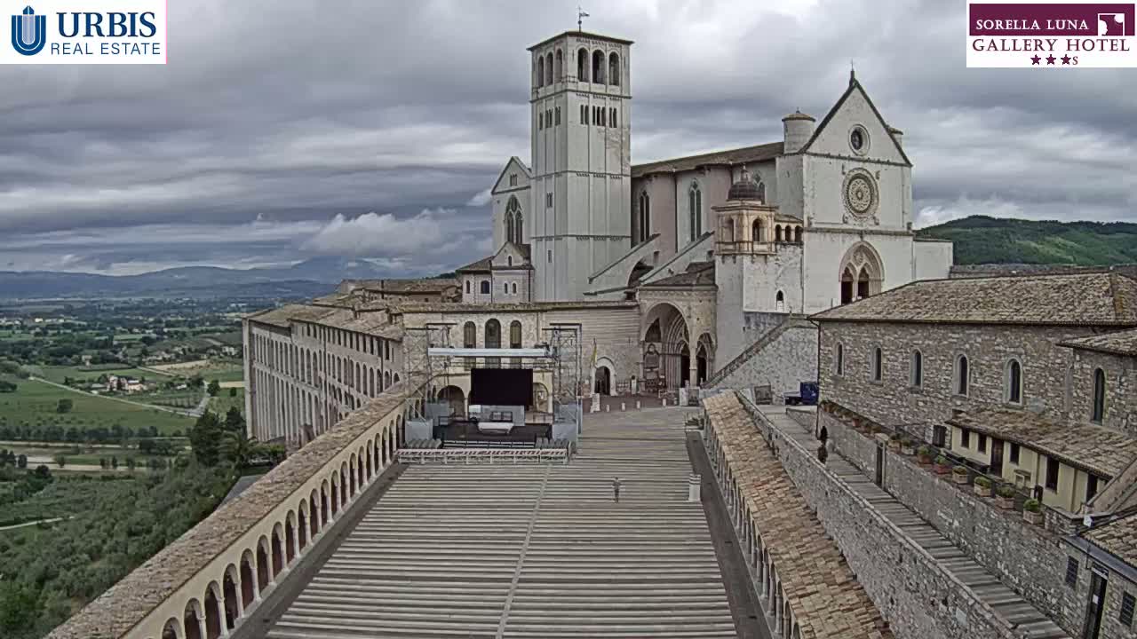 A large, light-colored basilica stands on a hill overlooking a valley under a cloudy sky, with a long, paved plaza stretching before it.