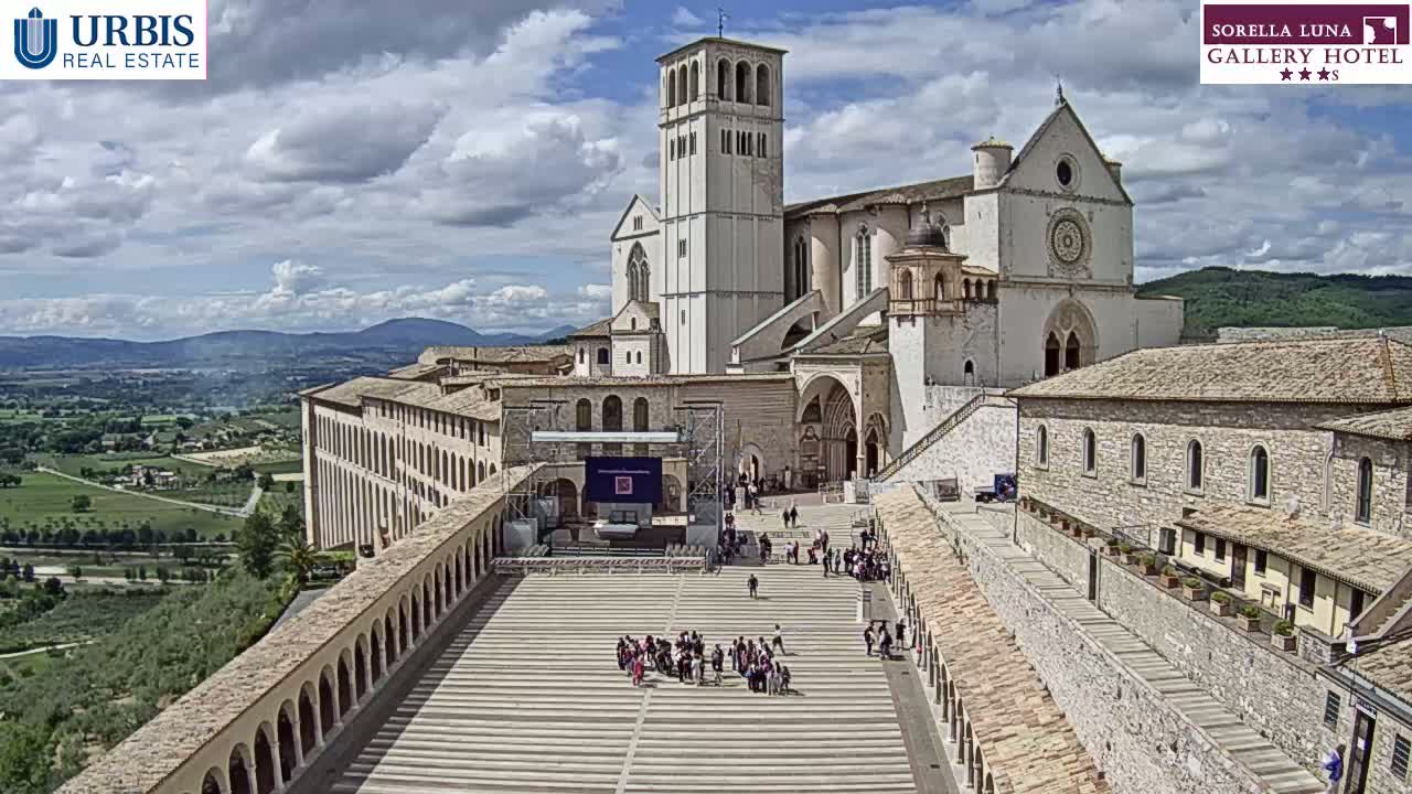 A large, light-colored basilica with a tall bell tower stands on a hill overlooking a valley under a partly cloudy sky, with a plaza below featuring several people.