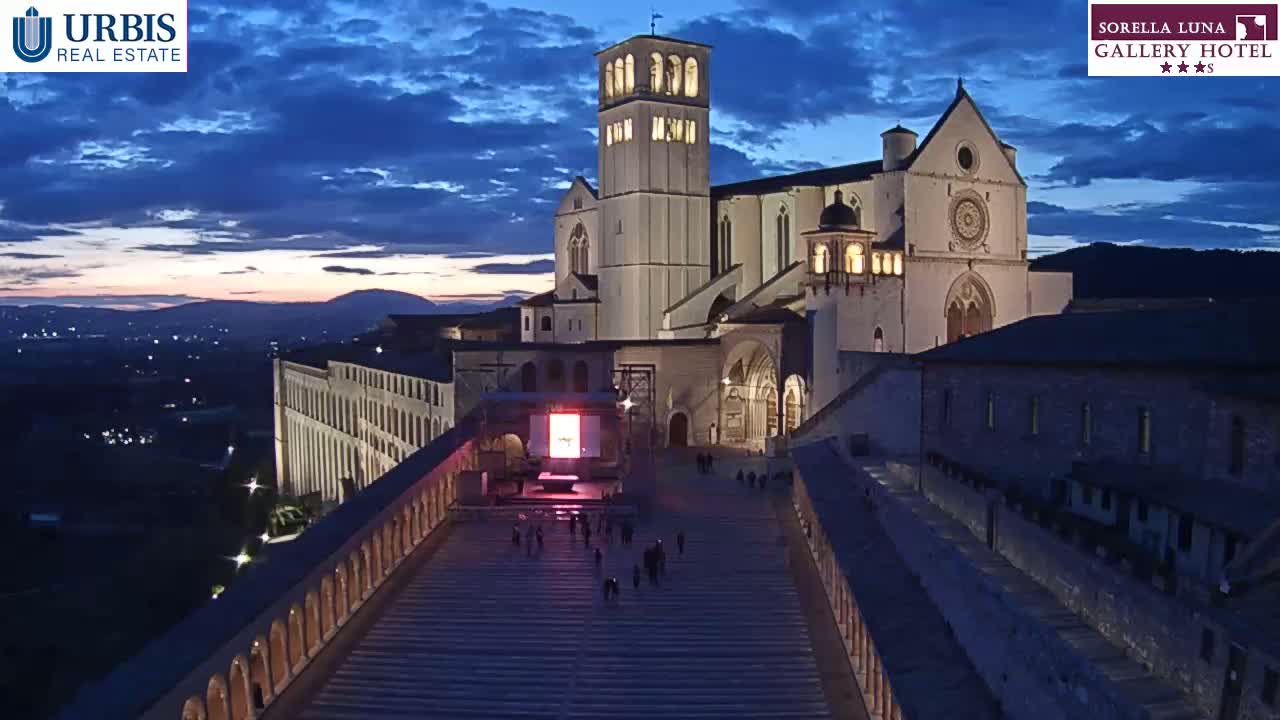 A large, illuminated basilica stands at twilight, overlooking a long, descending staircase and a city illuminated below under a partly cloudy sky.