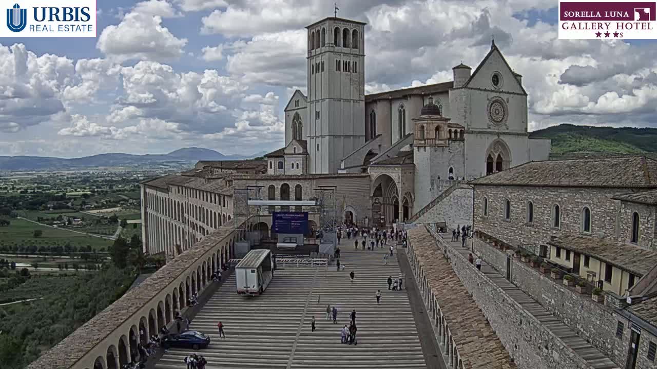 A large stone basilica and adjoining buildings are viewed from above on a partly cloudy day, with a plaza filled with people and a surrounding landscape visible in the distance.