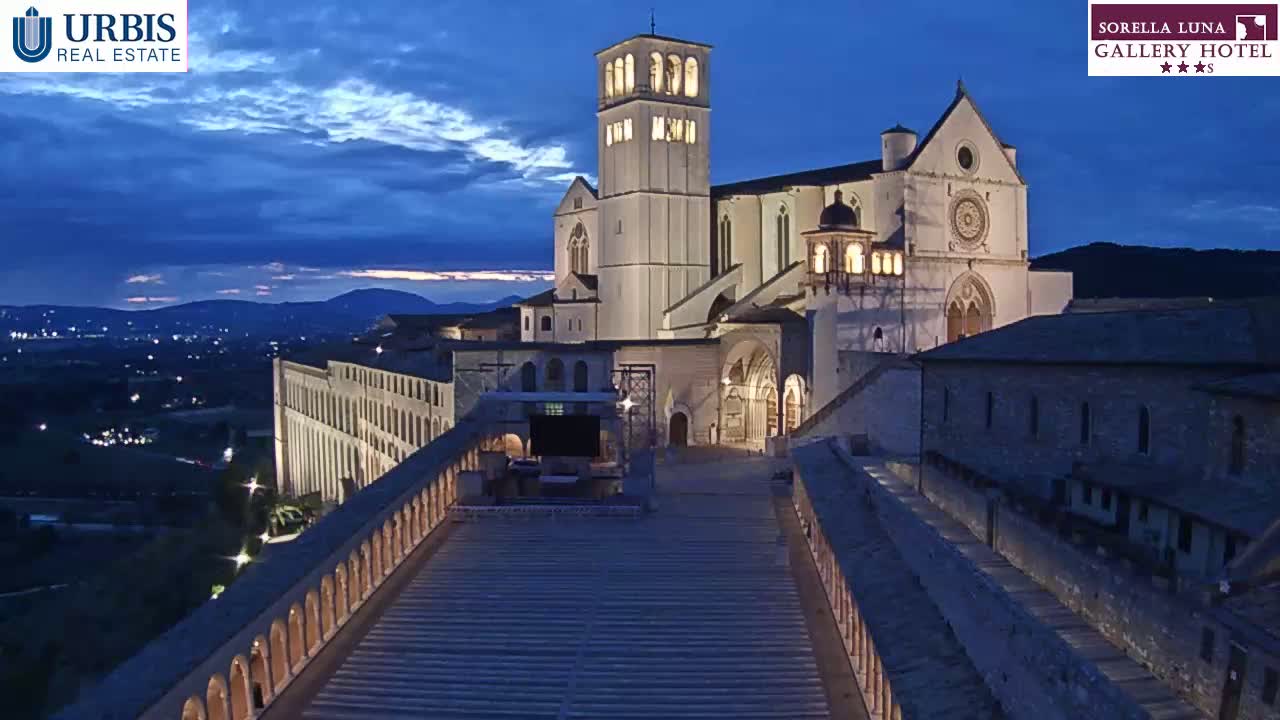 A large, illuminated basilica sits on a hilltop at twilight, overlooking a city at night, viewed from a long staircase leading to the building.