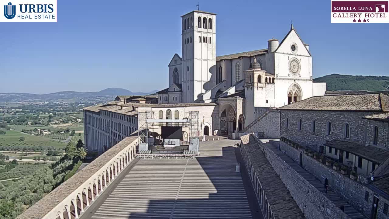 A large, light-colored stone basilica sits atop a hill overlooking a valley on a sunny day.