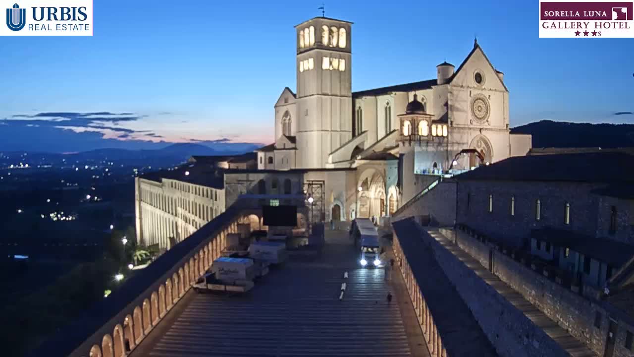A large, illuminated basilica stands on a hilltop at dusk, overlooking a city at the foot of the hill, with a long, colonnaded walkway leading down to it.