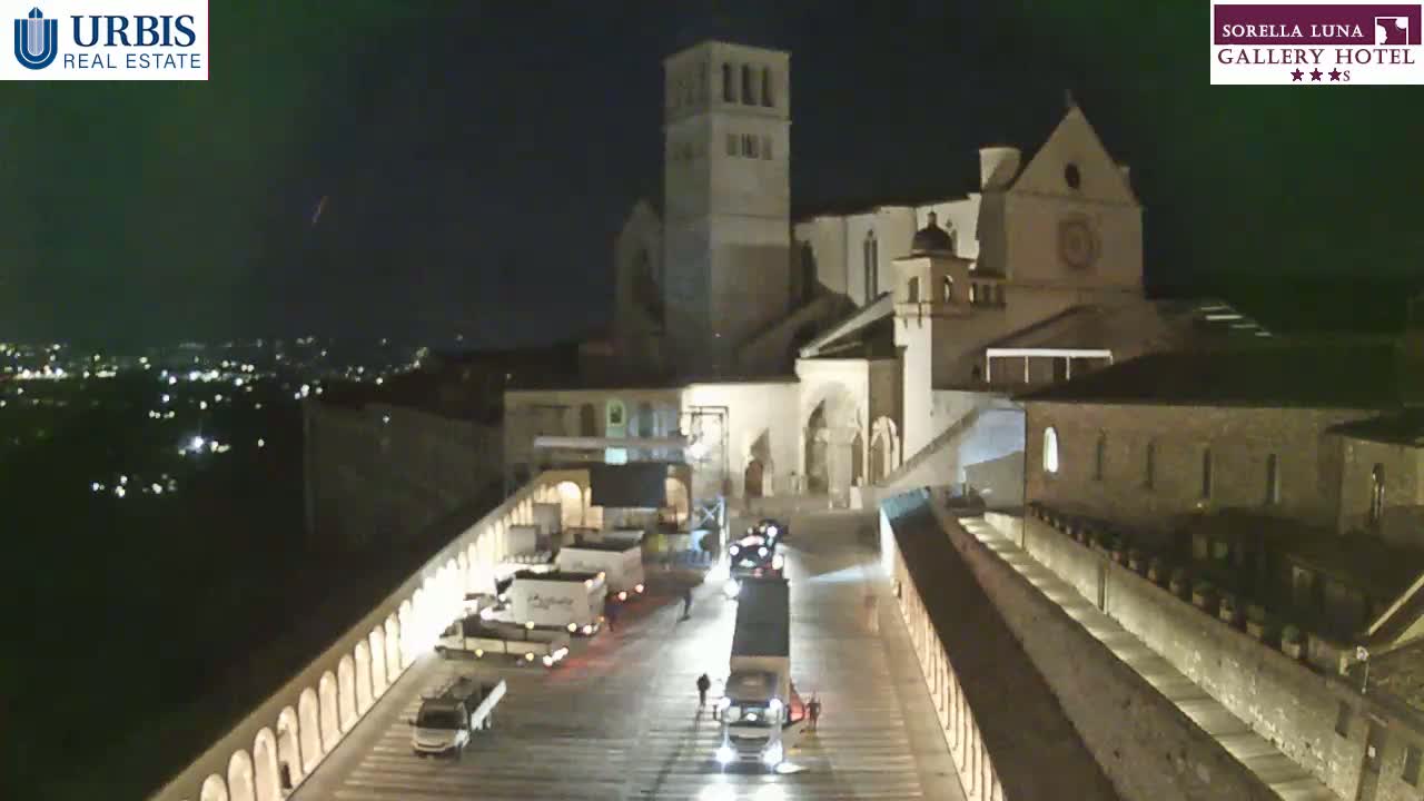 At night, a large, illuminated building complex is shown, with several trucks parked in a courtyard between two long, arched walls.