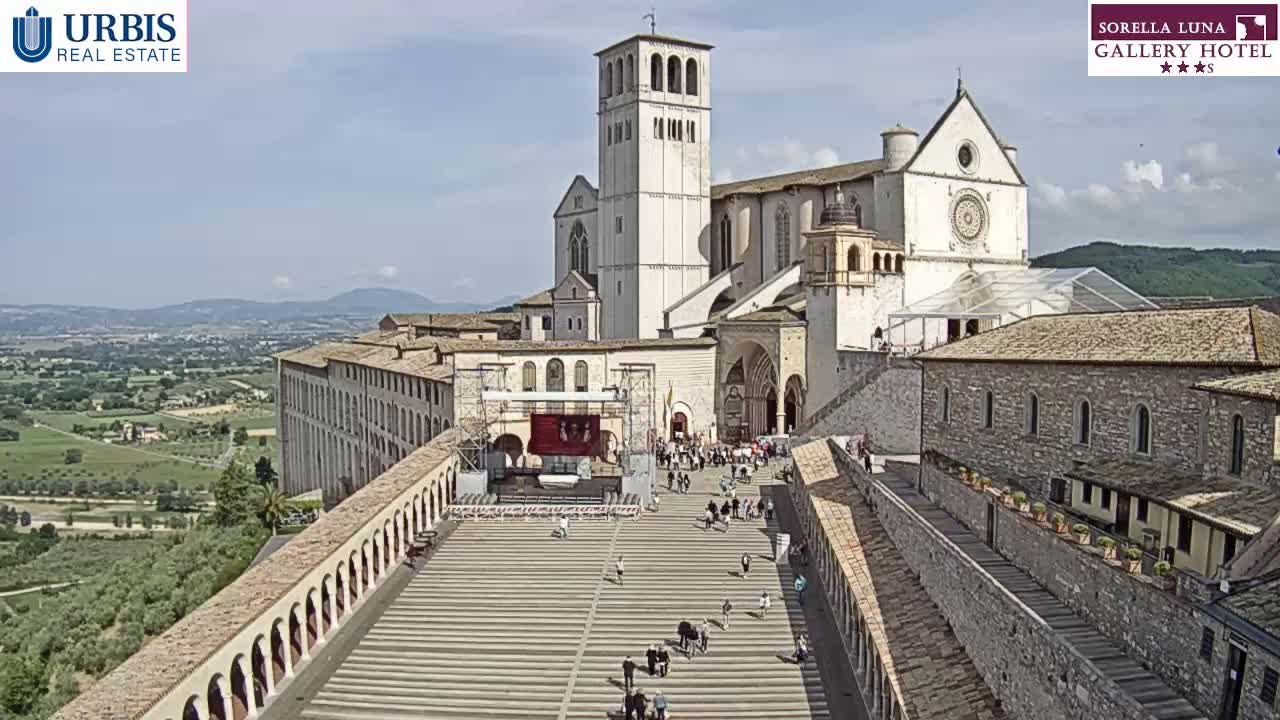 A large stone basilica and adjacent buildings are seen from above on a partly sunny day, with a paved plaza below containing a stage and a few people.