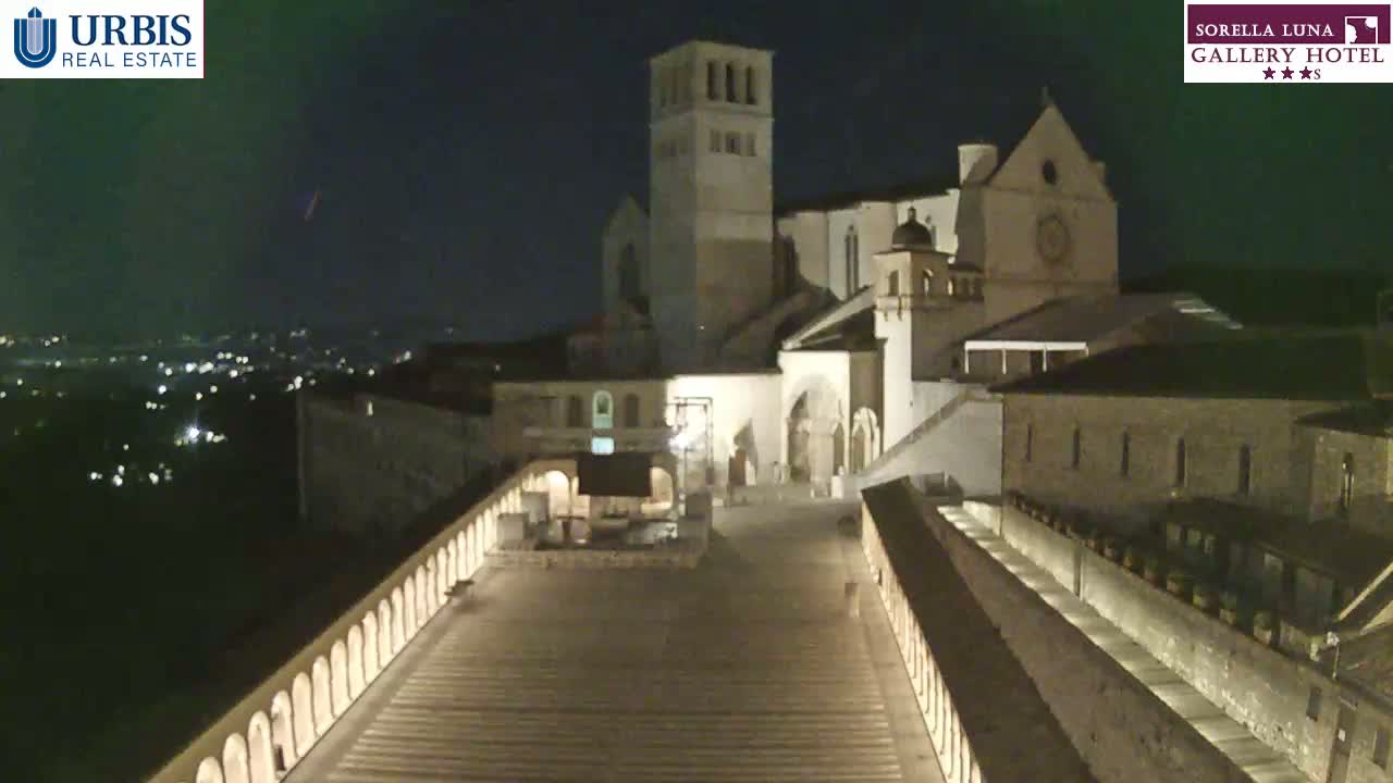 A large, illuminated stone structure, possibly a church, sits atop a hill at night, overlooking a city with a clear sky.