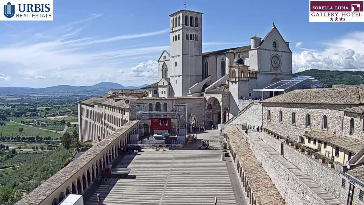 A large, light-colored stone basilica and adjacent buildings are situated on a hill overlooking a sprawling green valley under a mostly sunny sky.