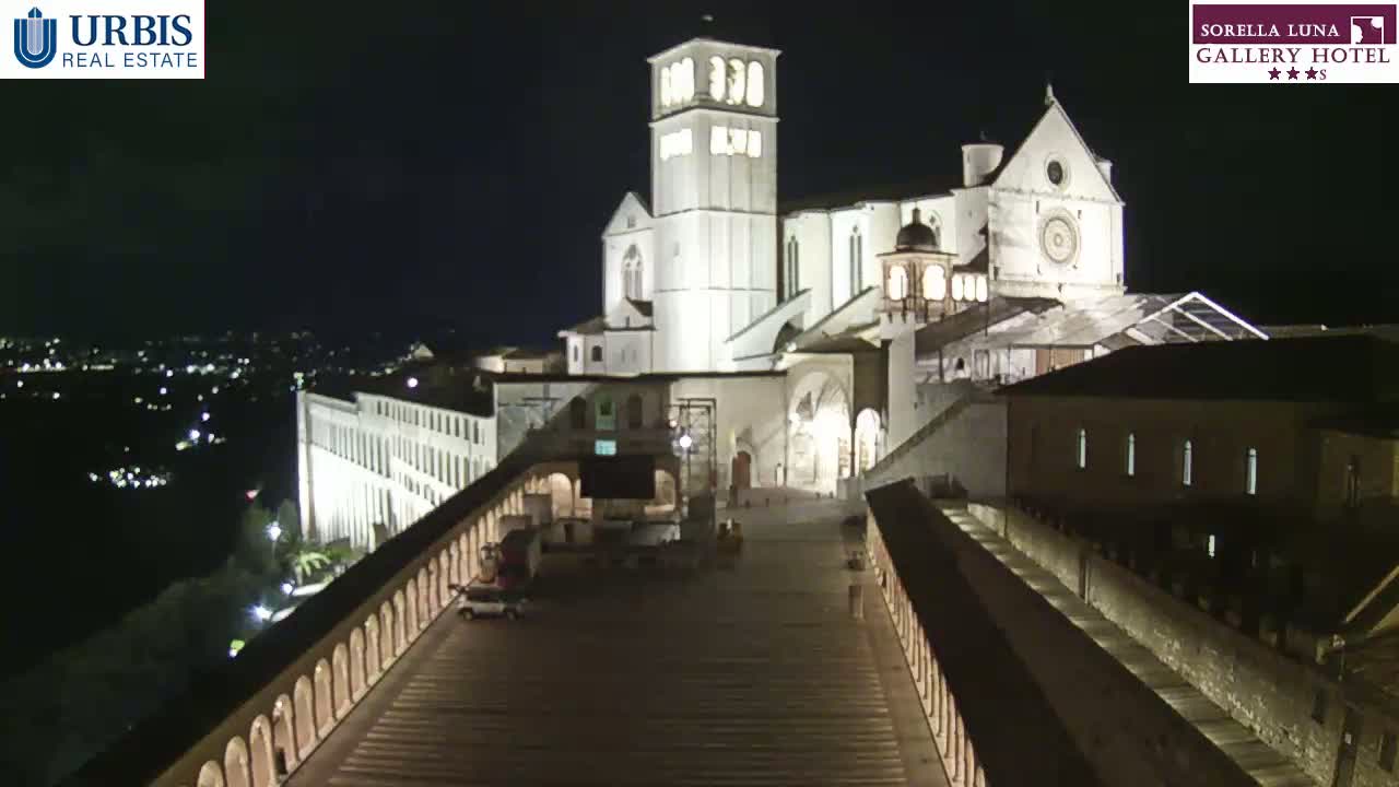 A large, illuminated basilica sits atop a hill at night, overlooked by a long, arched walkway.