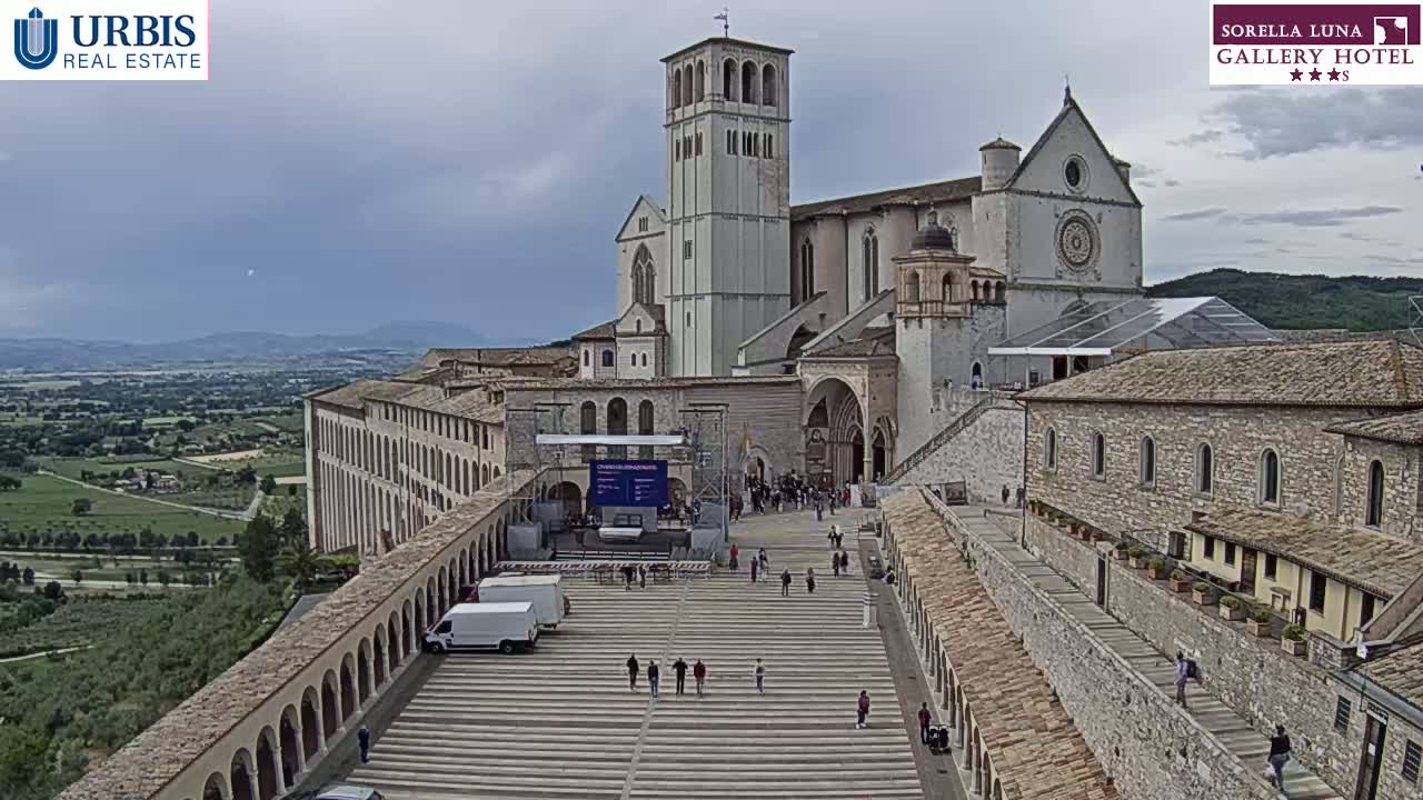 A large stone basilica and adjoining buildings are viewed from above on an overcast day, with a wide plaza in front containing several people and vehicles.