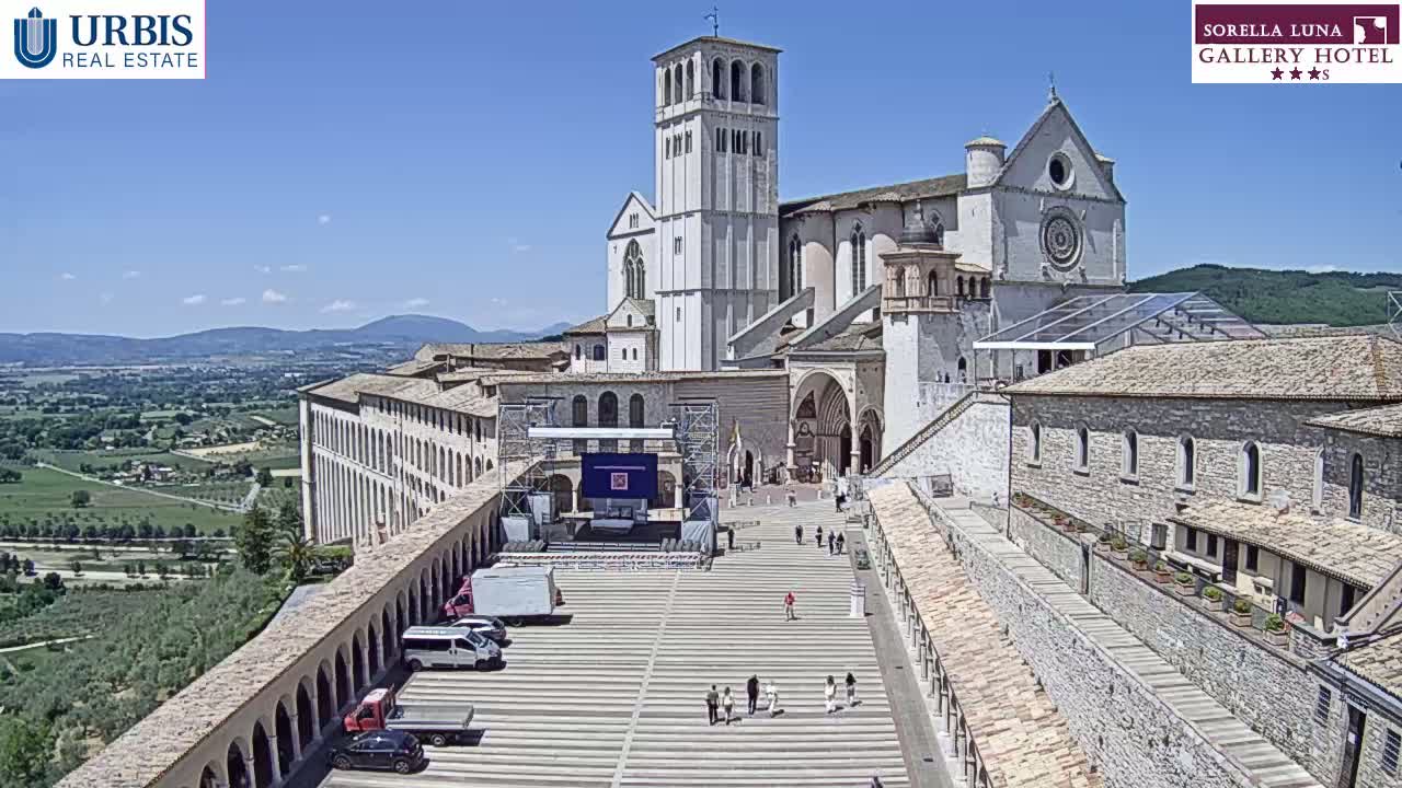 A large stone basilica and surrounding buildings are seen on a sunny day, with a paved plaza in front and a view of rolling hills in the distance.