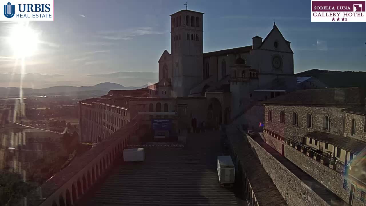 A large stone basilica stands on a hilltop overlooking a town, bathed in the warm light of a setting sun.