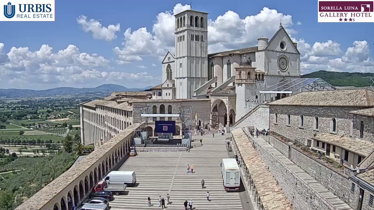 A large stone basilica sits on a hill overlooking a valley under a partly cloudy sky.