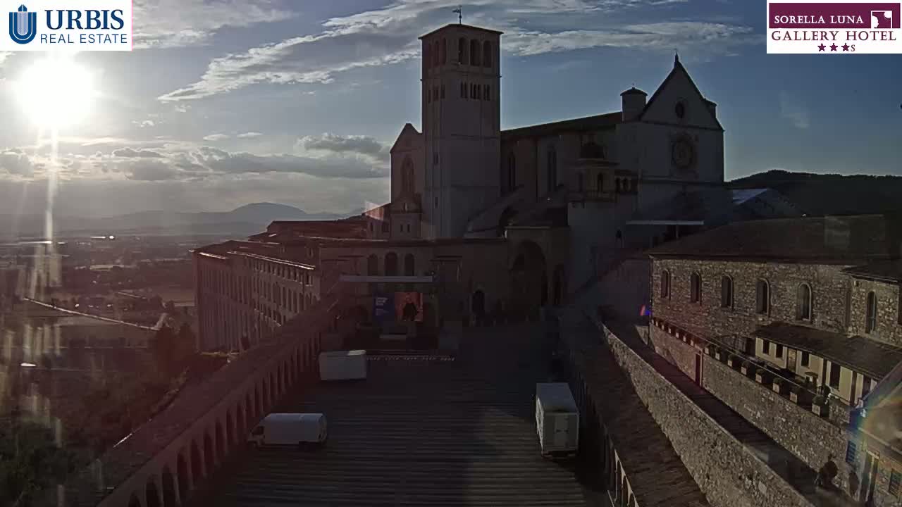 A large stone building, possibly a church or monastery, sits on a hill overlooking a town under a partly sunny sky.
