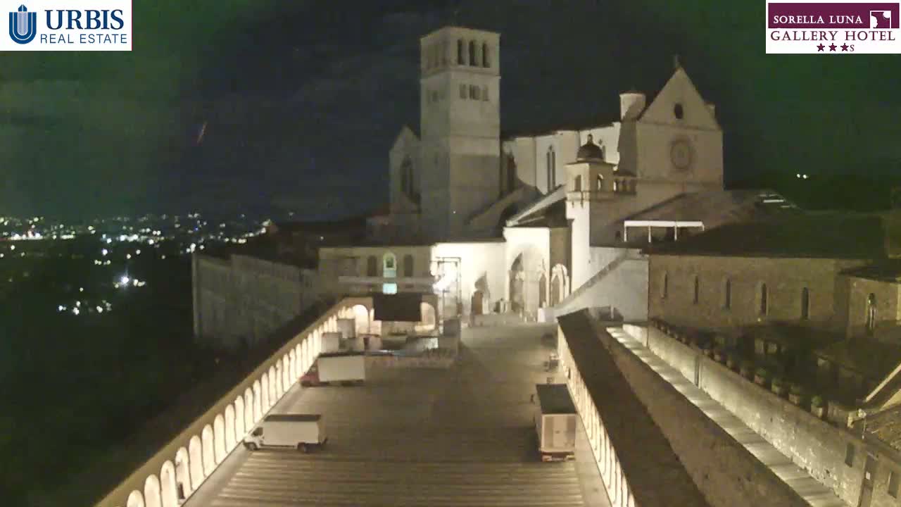 A large, illuminated stone building complex is seen at night, with a few vehicles parked in a plaza below, and city lights visible in the distance.