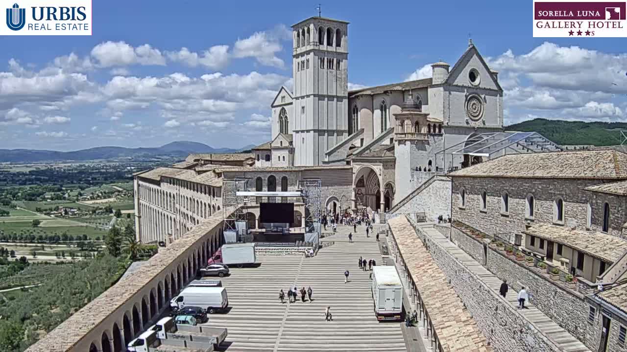 A large stone basilica and adjoining buildings stand on a hill overlooking a valley on a partly cloudy day, with a plaza in front containing several vehicles and a few people.