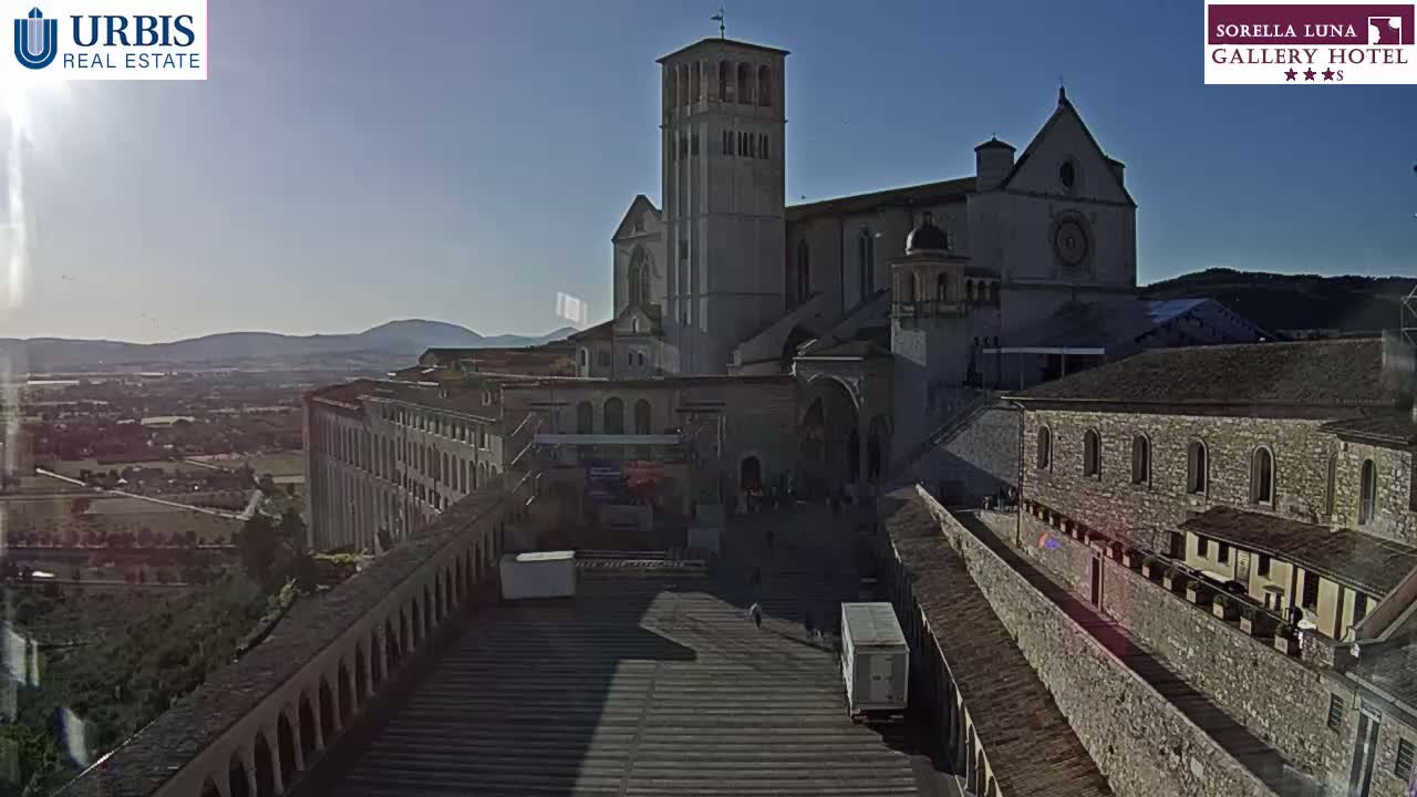 A large stone basilica and adjacent buildings are situated on a hill overlooking a valley under a clear, sunny sky.