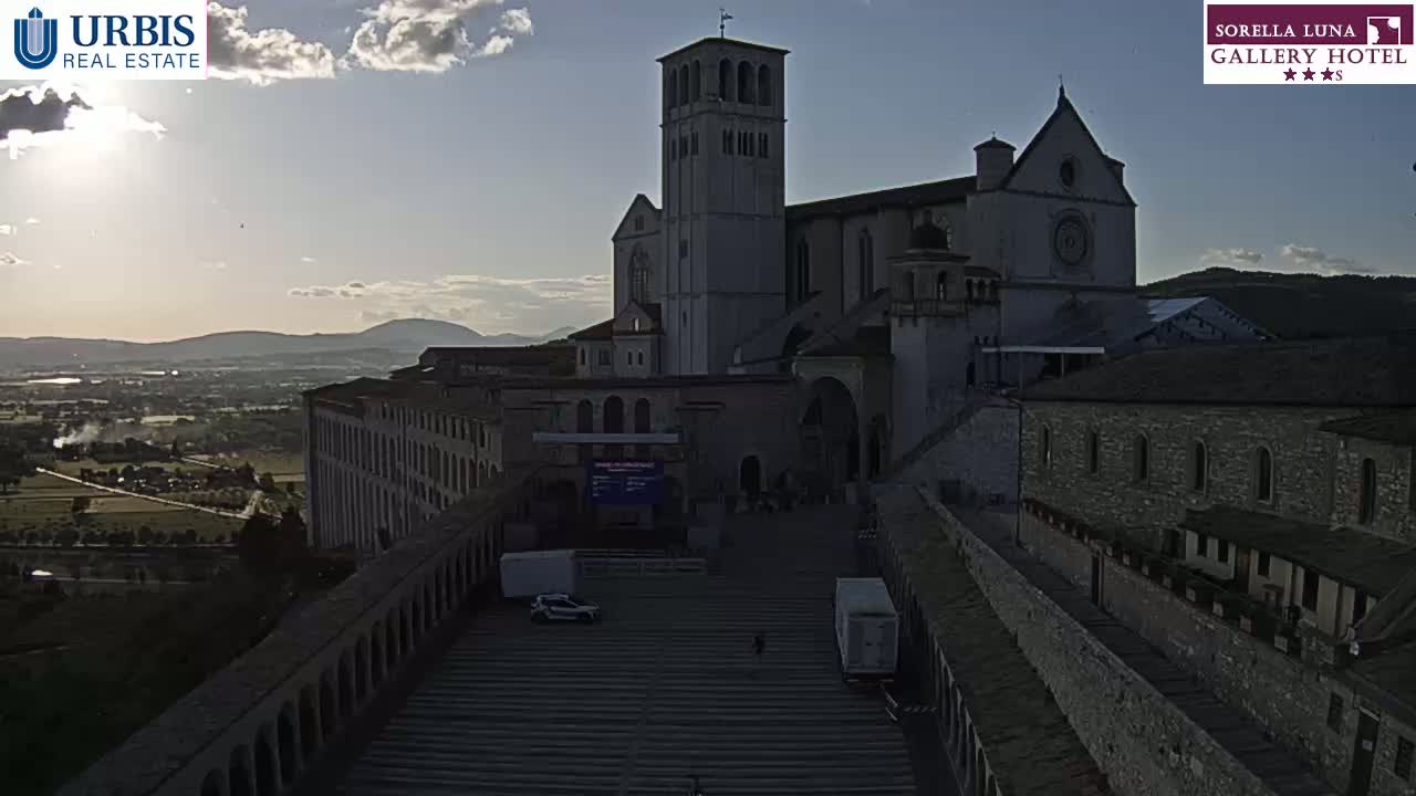 A large stone basilica sits on a hill overlooking a valley under a partly cloudy sky.