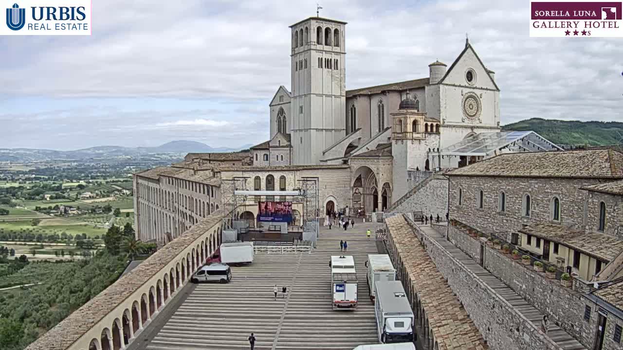 An expansive view of a large stone basilica and adjacent buildings on a slightly overcast day, with a valley and distant hills visible in the background.