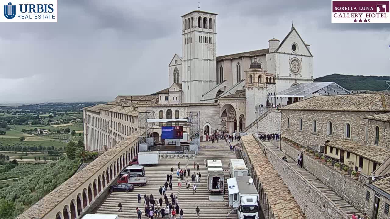 A large stone basilica and adjoining buildings are visible on a cloudy day, with a crowd of people gathered in the plaza below and various vehicles parked nearby.