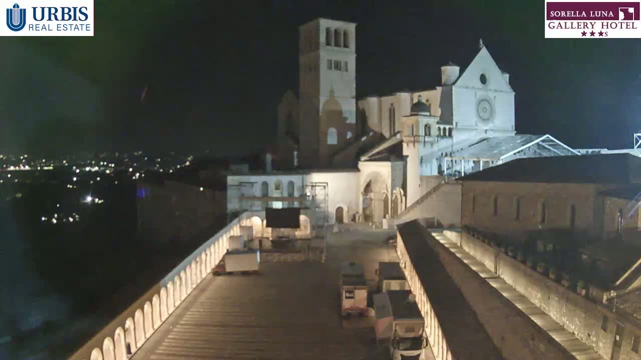 A nighttime view of a large stone building complex, possibly a church or monastery, with a paved area and several vehicles in the foreground.