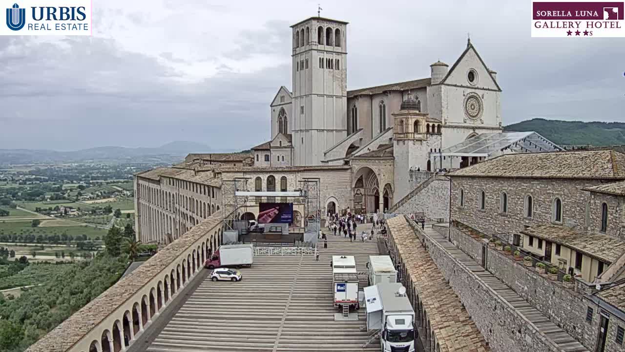 A large stone basilica overlooks a plaza with several vehicles and a temporary stage, all under a cloudy sky.
