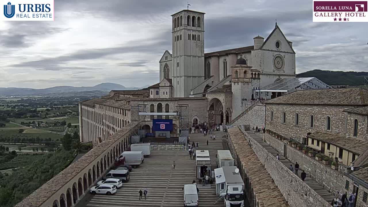 A large stone basilica and adjacent buildings are seen on an overcast day, with a plaza in front containing several vehicles and a few people.