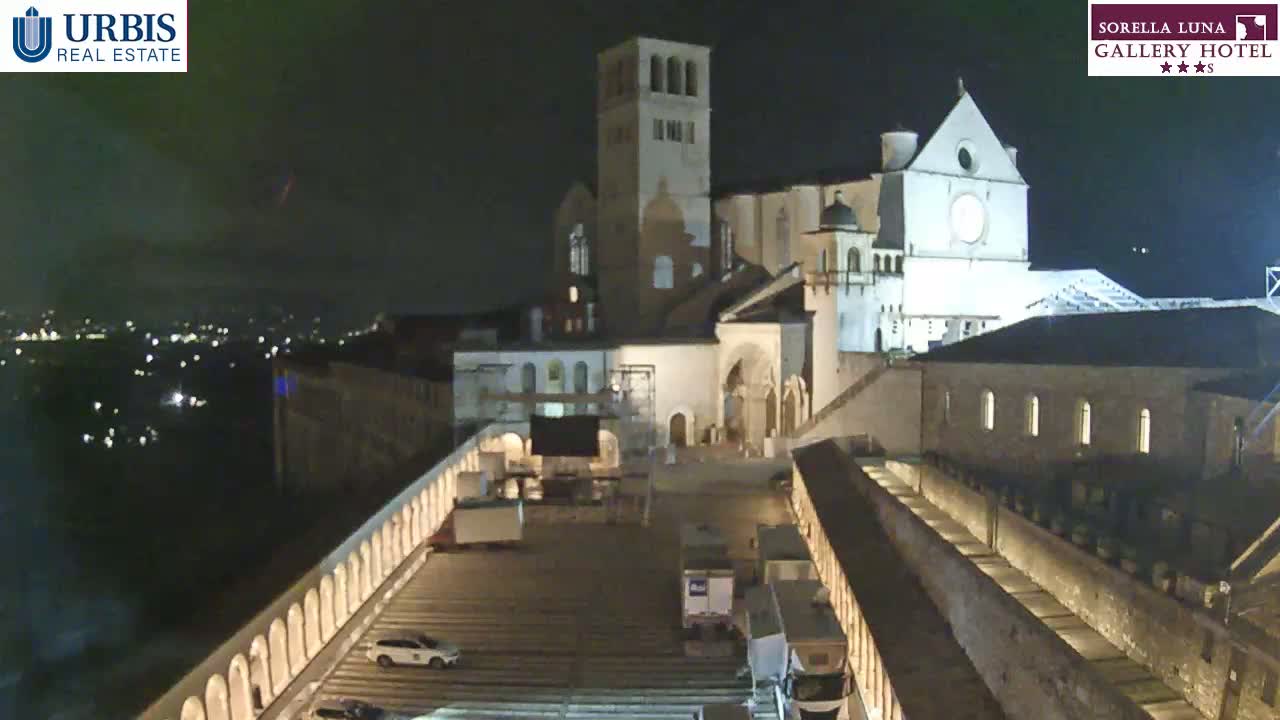 A large, illuminated church is seen at night, with a few vehicles parked in a plaza below, and a city skyline visible in the distance.