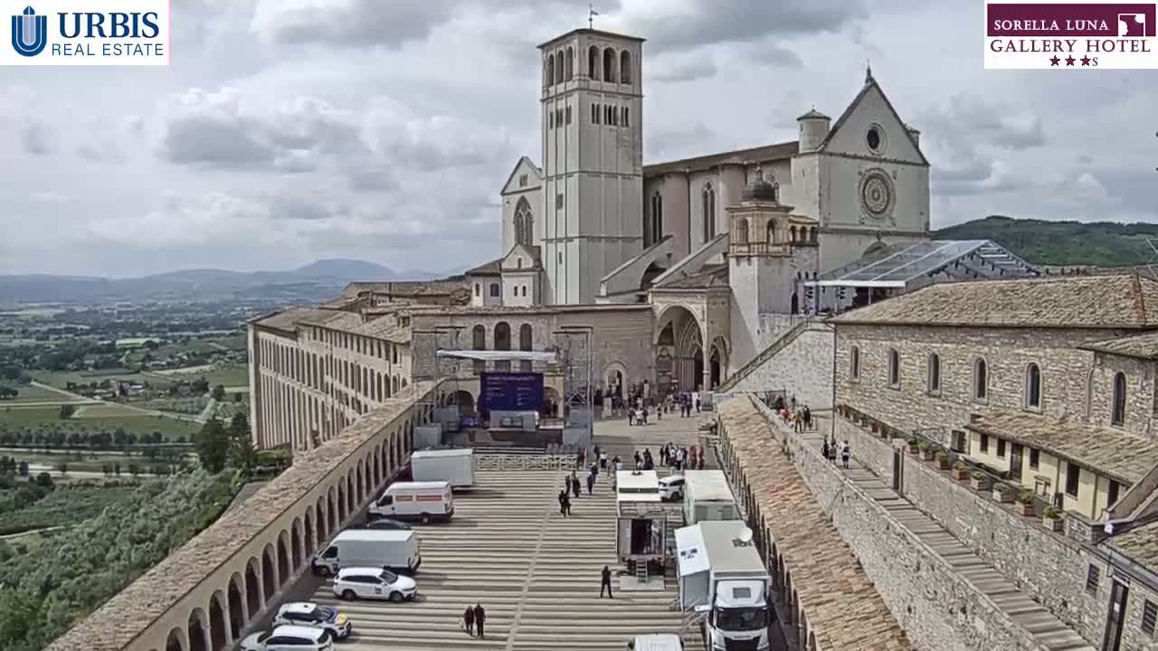 A large stone basilica and adjacent buildings are seen from above on a partly cloudy day, with a square in front containing several vehicles and a few people.