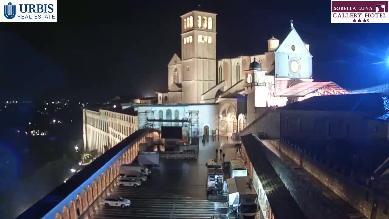 A large, illuminated building complex, possibly a church or cathedral, is seen at night, with several vehicles parked in a plaza below.
