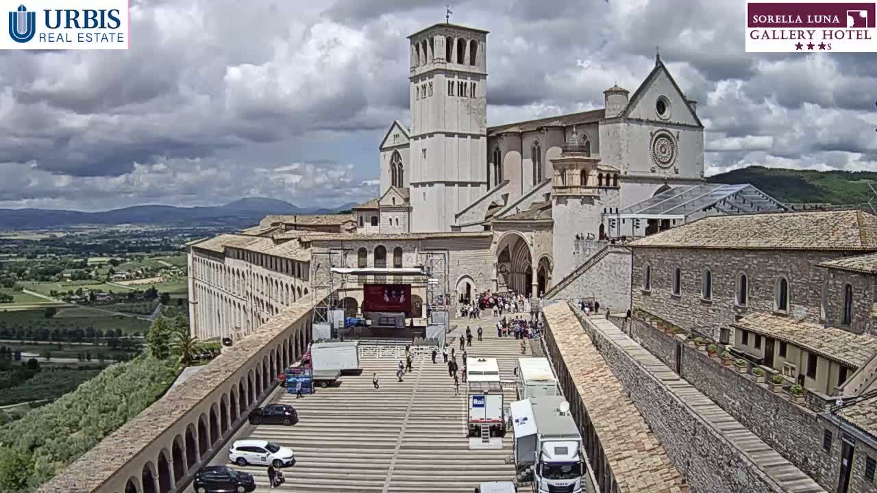 A large stone basilica and adjacent buildings are situated on a hill overlooking a valley under a partly cloudy sky.