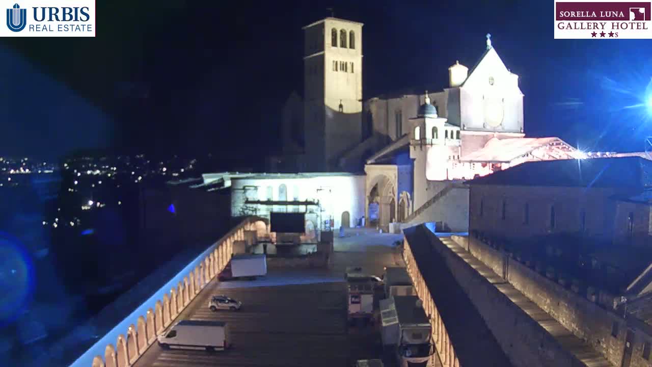 A nighttime aerial view of a large, illuminated stone building complex, possibly a church or monastery, with a plaza below containing several vehicles and surrounded by walls with arched walkways.