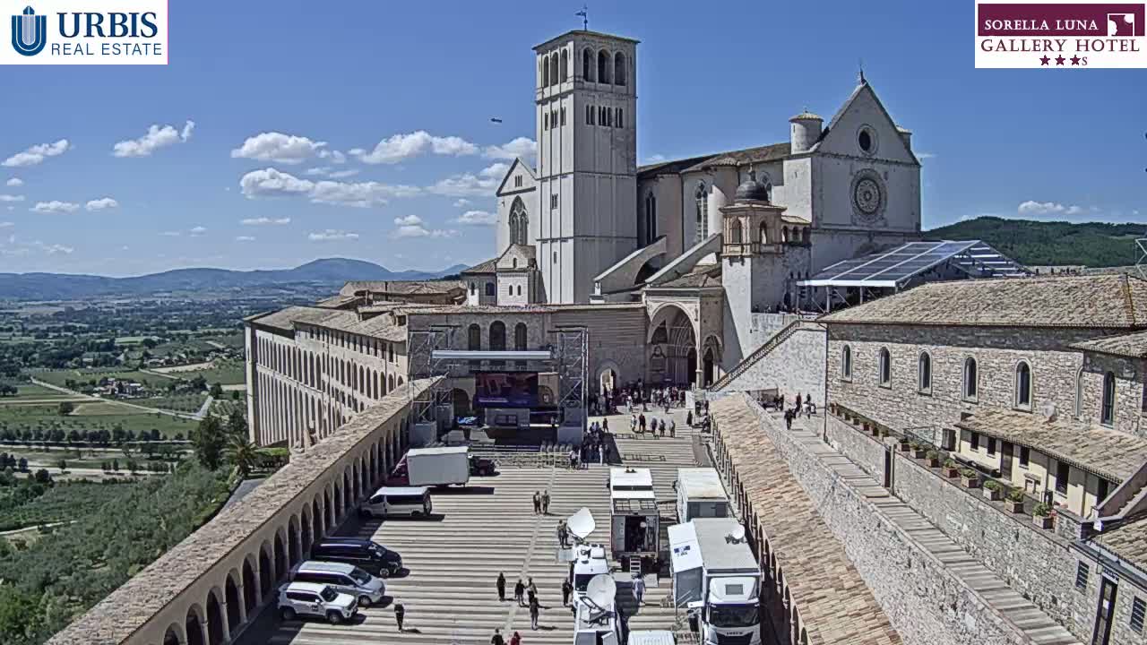 A large stone basilica and adjacent buildings are seen on a sunny day, with a plaza in the foreground containing several vehicles and a few people.