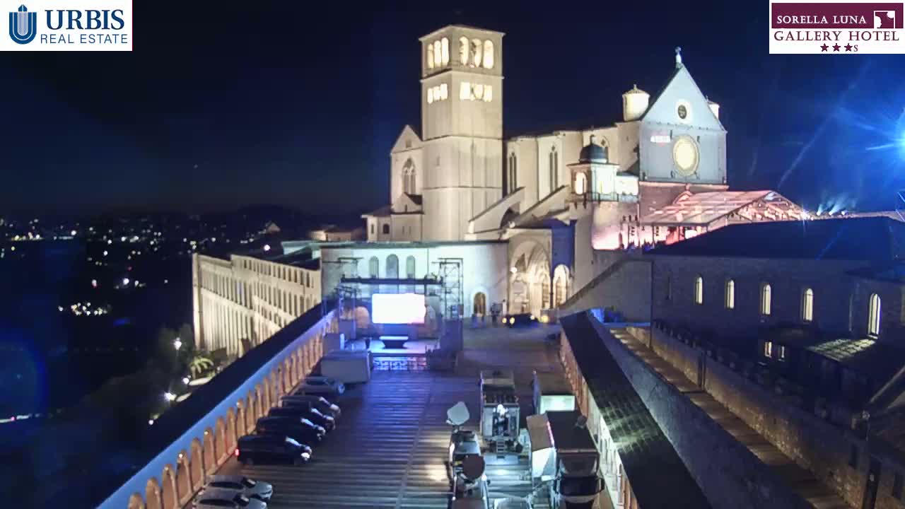 A large, illuminated building complex, possibly a church, at night, with a parking lot containing several vehicles and broadcasting equipment in the foreground.