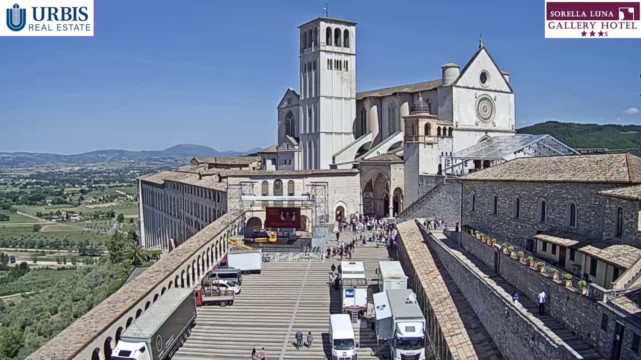 A large stone basilica and adjacent buildings are viewed from above on a sunny day, with a plaza below containing several trucks and a small gathering of people.
