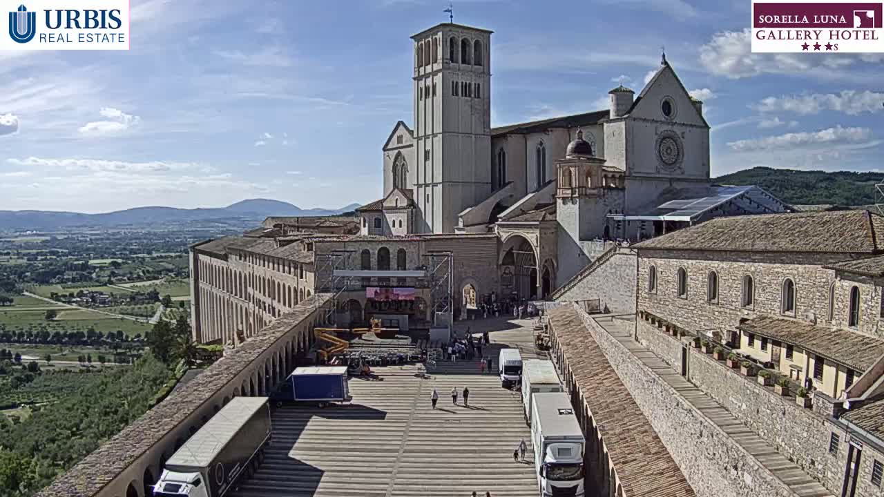 A large stone basilica sits atop a hill overlooking a valley under a mostly sunny sky, with several trucks parked in a plaza in front of it.