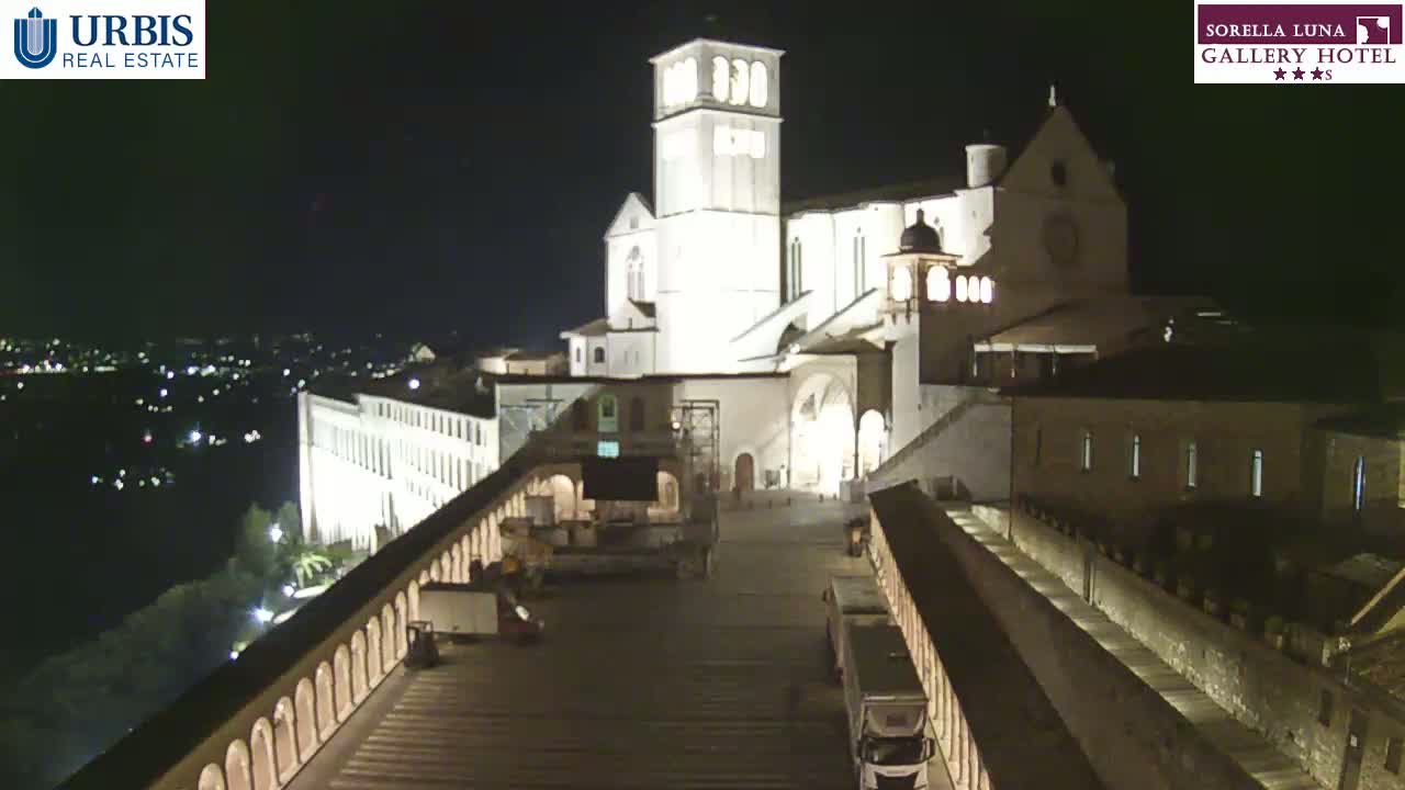 A large, illuminated basilica stands at night, overlooking a city, with several vehicles parked on a long, paved pathway below.