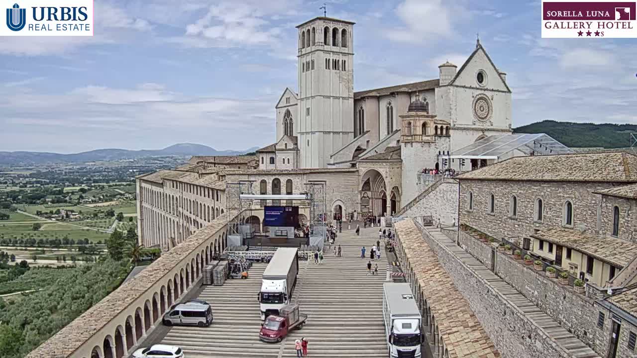 A large stone basilica and adjoining buildings are seen from an elevated viewpoint on a partly cloudy day, with a wide plaza in front featuring several trucks and a small gathering of people.