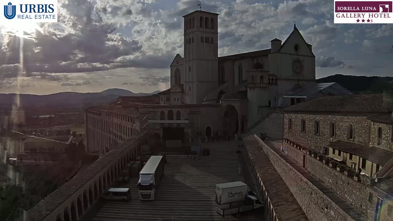 A large stone basilica and surrounding buildings are viewed from above under a partly cloudy sky, with several delivery trucks parked in a plaza below.